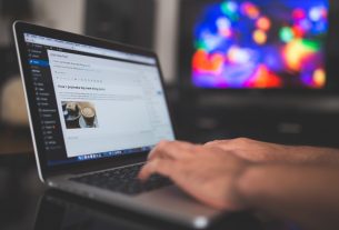 Hands typing on a laptop with a blog post visible, cozy indoor setting with colorful screen in background.