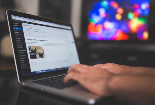 Hands typing on a laptop with a blog post visible, cozy indoor setting with colorful screen in background.